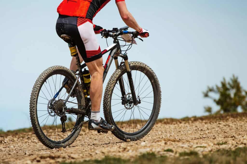 Een persoon in een rood en zwart fietstenue rijdt op een mountainbike op een rotsachtig zandpad op Aruba. Met dikke, modderige banden en waterflessen aan het frame suggereert de scène een spannend buitenavontuur.
