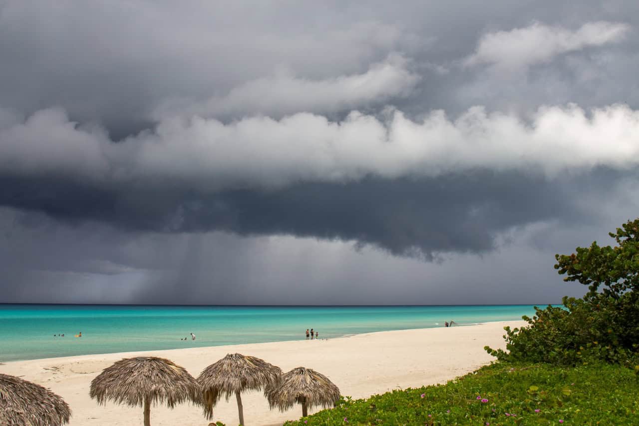 A beach of white sand, turquoise waters, and thatched umbrellas lies under a dramatic sky as dark storm clouds approach – typical of Bonaire's rainy season. A few people are walking along the shoreline, bordered by lush green plants.