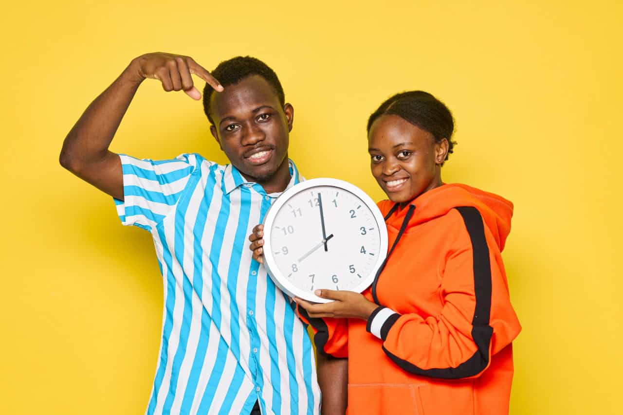 Two smiling people stand in front of a yellow background. One person in a striped shirt points to a wall clock held by the other person in an orange hoodie. On the clock it is 7:05 am.