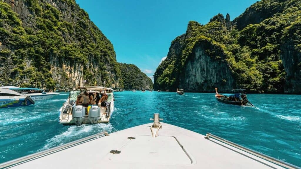 Vista desde la proa de un barco que navega por aguas azules y transparentes entre acantilados de piedra caliza escarpados y frondosos, con varios otros barcos y personas visibles disfrutando de la pintoresca laguna tropical.