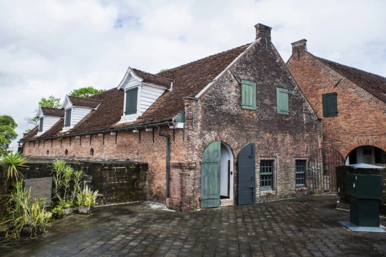 Een historisch bakstenen gebouw in Fort Zeelandia in Suriname, met steile daken bedekt met dakspanen en groene houten luiken, staat op een binnenplaats van natte stenen. In de schaduw van een stenen muur groeien weelderige planten onder een bewolkte hemel.