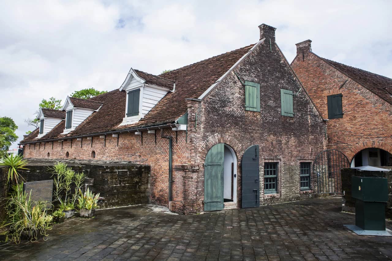 Een historisch bakstenen gebouw in Fort Zeelandia in Suriname, met steile daken bedekt met dakspanen en groene houten luiken, staat op een binnenplaats van natte stenen. In de schaduw van een stenen muur groeien weelderige planten onder een bewolkte hemel.