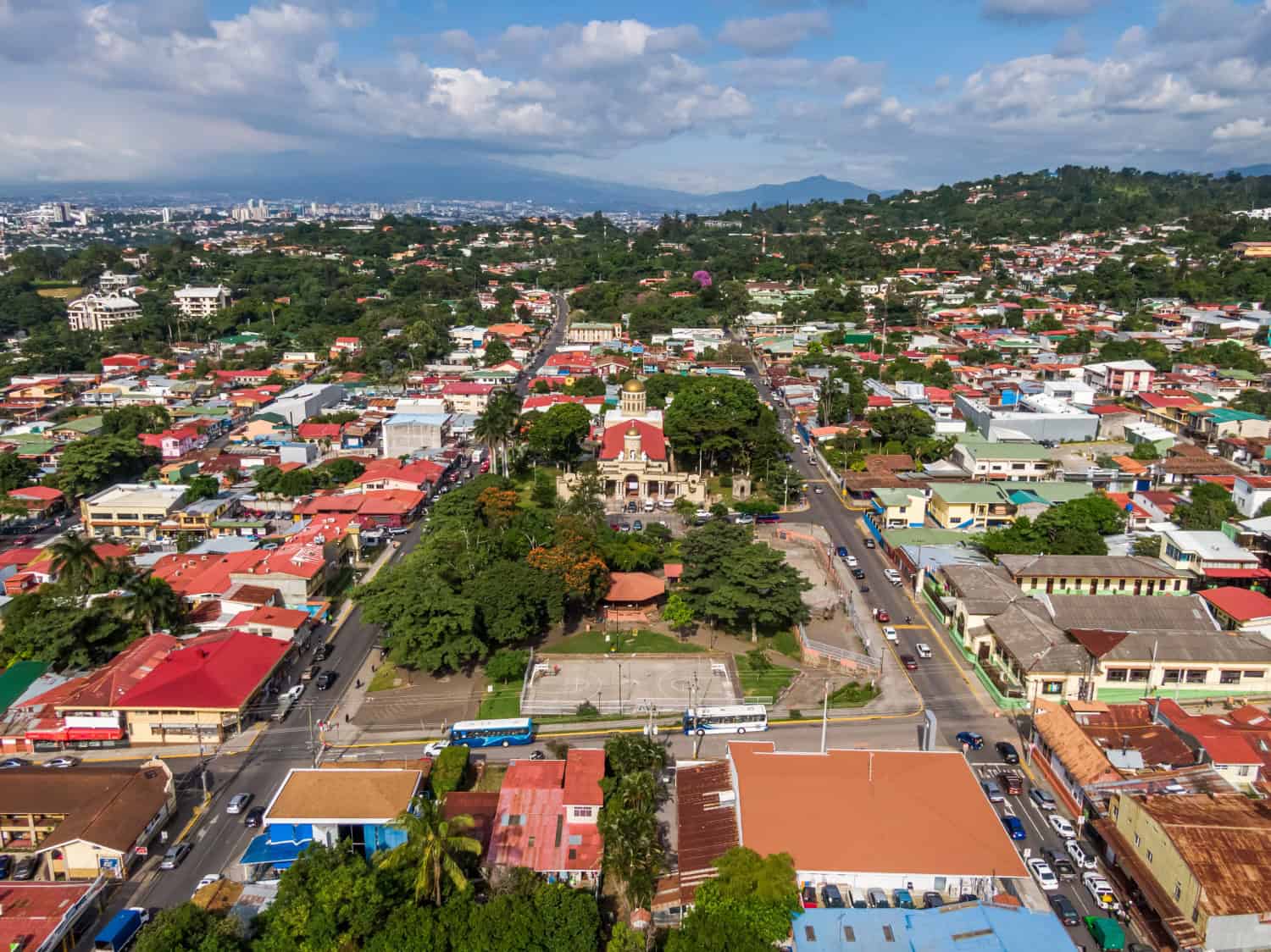 Luchtfoto van het stadsplein van Paramaribo, omringd door kleurrijke gebouwen en straten met auto's en bussen; verre heuvels en bergen onder een deels bewolkte lucht vormen de achtergrond. Weelderige bomen vullen het park in het midden van de afbeelding.