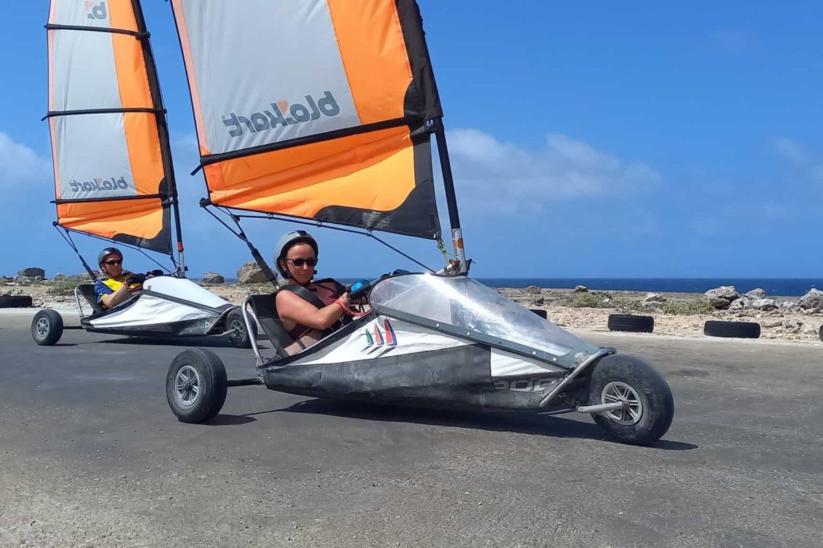 Two children ride a Bonaire land sailing double kart for ages 3 to 8, featuring orange and white sails, on a paved oceanfront road under a bright blue sky, with rocky shoreline and tires outlining the course in the background.