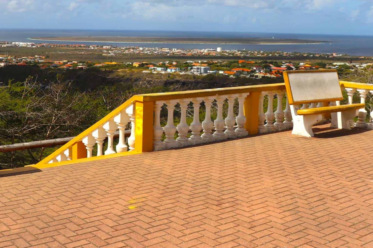 A yellow and white balcony with a bench overlooks Bonaire's coast on the Private North Tour, featuring colorful rooftops, green hills, and the distant ocean beneath a partly cloudy sky.