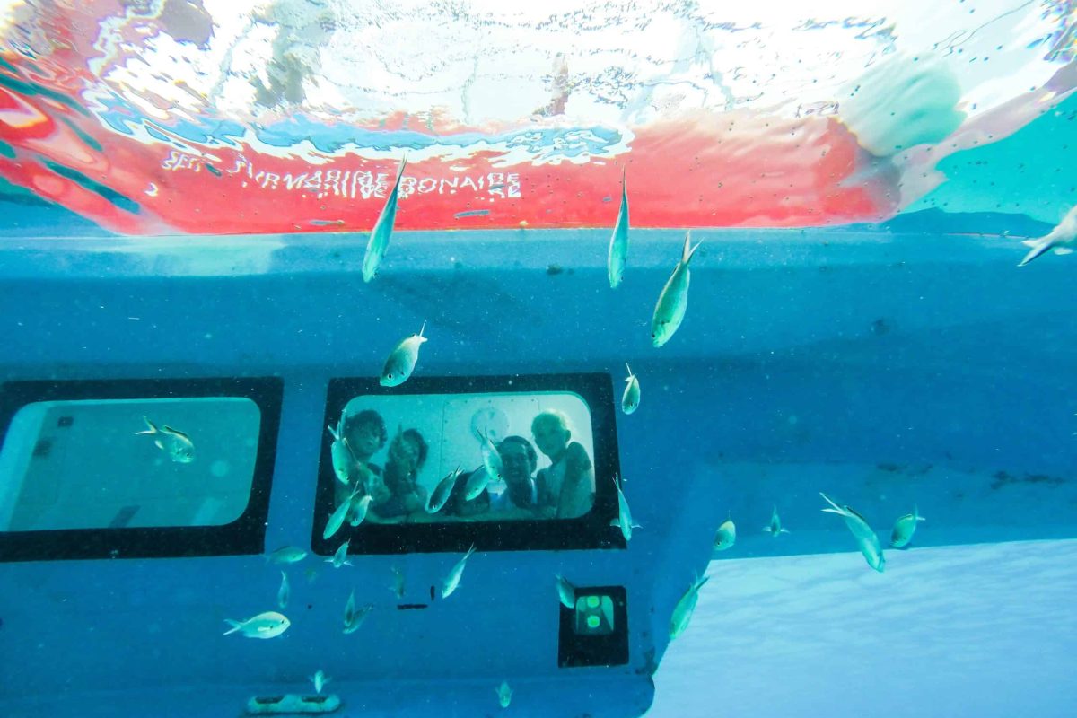 Underwater view of the Semi Submarine (Bonaire) with people looking out the windows, surrounded by small fish in clear blue water. Red letters are seen on the surface above the submarine.