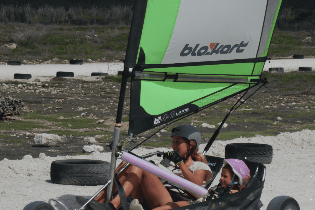 An adult steers a Land sailing double kart for children 3 to 8 years (Bonaire) with a child passenger, both wearing helmets, as they ride on a dirt track bordered by tires and grass.