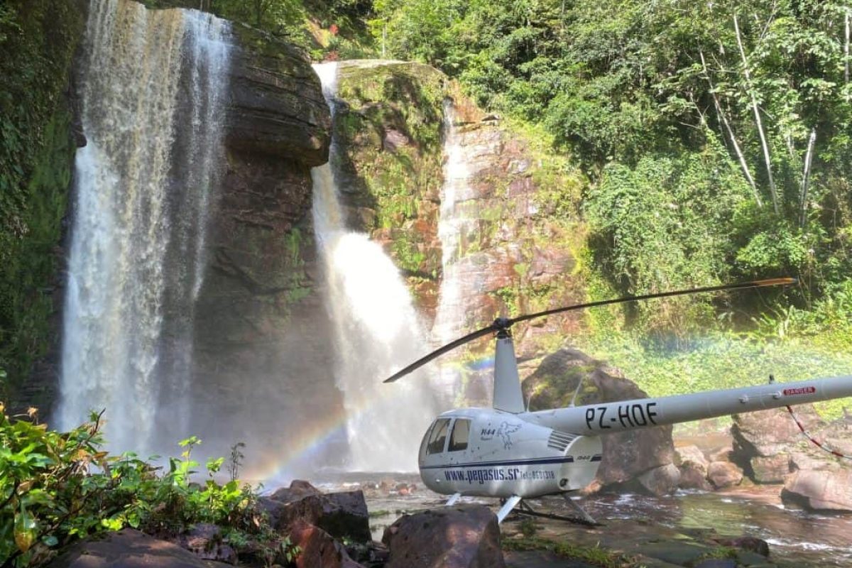 Un helicóptero blanco para el "Vuelo en helicóptero sobre (Surinam)" está aparcado sobre unas rocas cerca de una exuberante cascada, donde la luz del sol forma un pequeño arco iris en la niebla rodeada de vegetación.