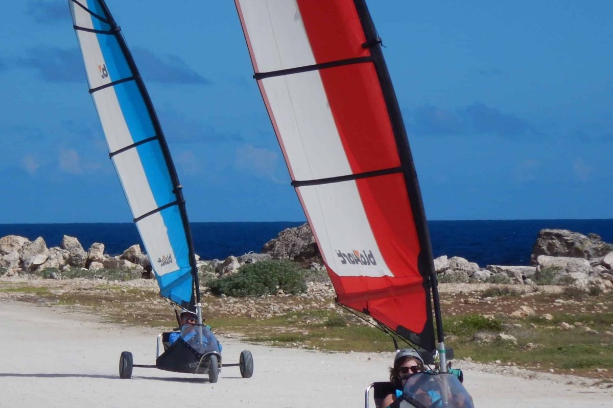 Twee mensen rijden op Landzeilen 50 minuten (Bonaire) landzeilen met blauwwitte en roodwitte zeilen over een zandpad langs zee, omringd door rotsen onder een helderblauwe hemel.