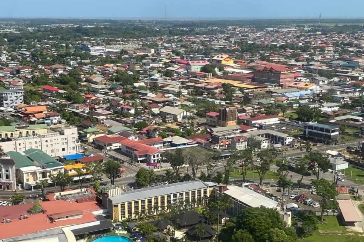 Vista aérea durante un Vuelo en helicóptero sobre Surinam, que muestra edificios de poca altura, tejados de colores, calles arboladas y una piscina enmarcada por vegetación bajo un cielo azul despejado.