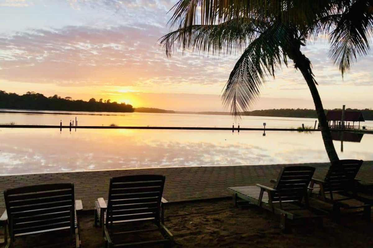 Ligstoelen aan de rivieroever bij zonsondergang, met een palm op de voorgrond