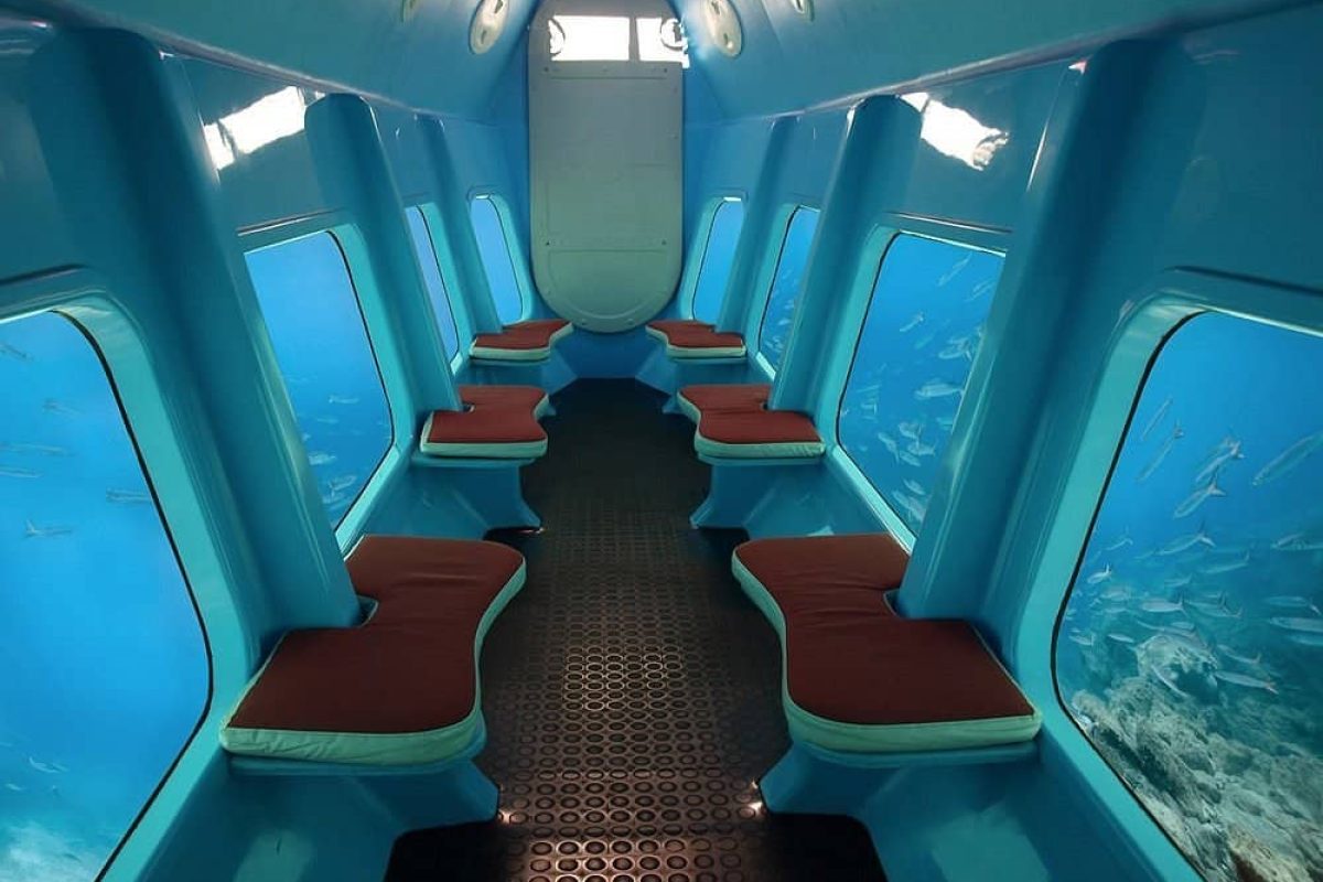 The Semi Submarine (Bonaire) features a blue interior with large side windows, red-cushioned bench seats, and offers views of fish swimming outside in the ocean.