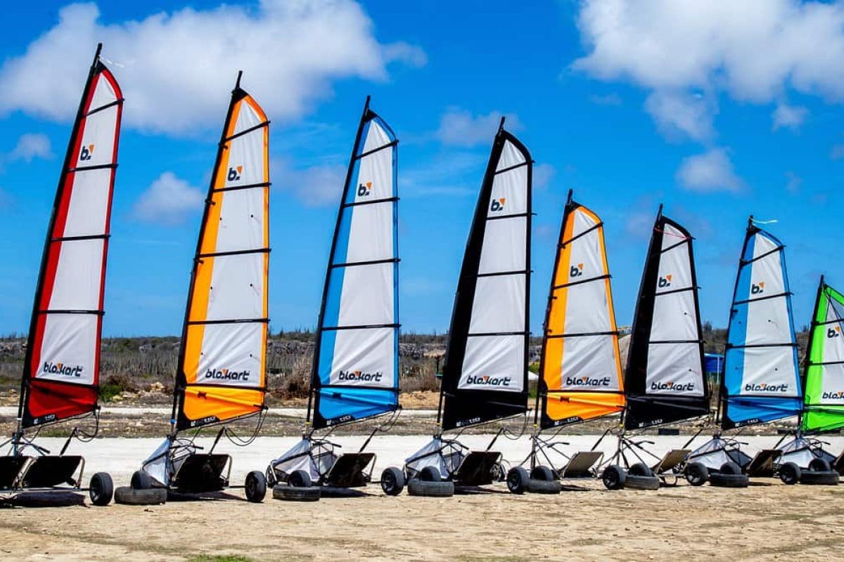 Several Land sailing double karts for children aged 3 to 8 years (Bonaire), featuring large red, orange, white, blue, and yellow sails, are lined up on a sunny sandy beach beneath a bright blue sky with scattered clouds.